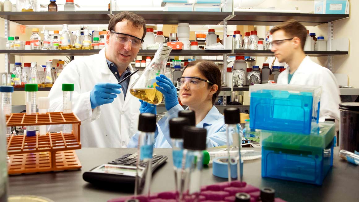Bryant Professor Chris Reid points to a glass flask with some students in a lab.