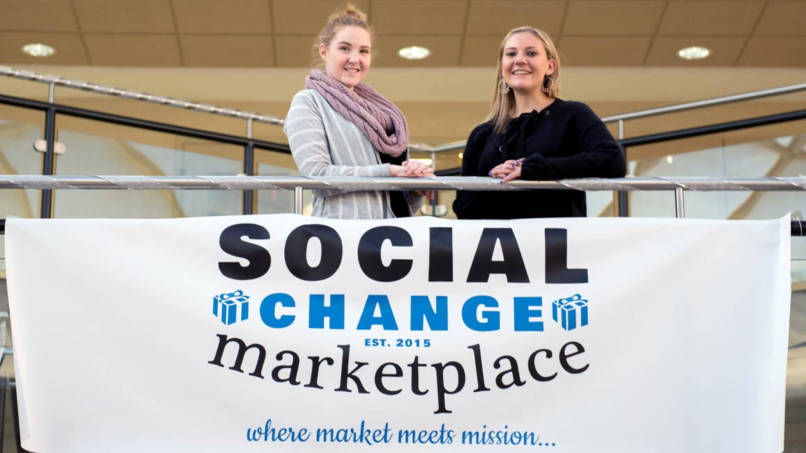 Two students pose in front of a Social Change Marketplace banner inside the Rotunda at Bryant University.