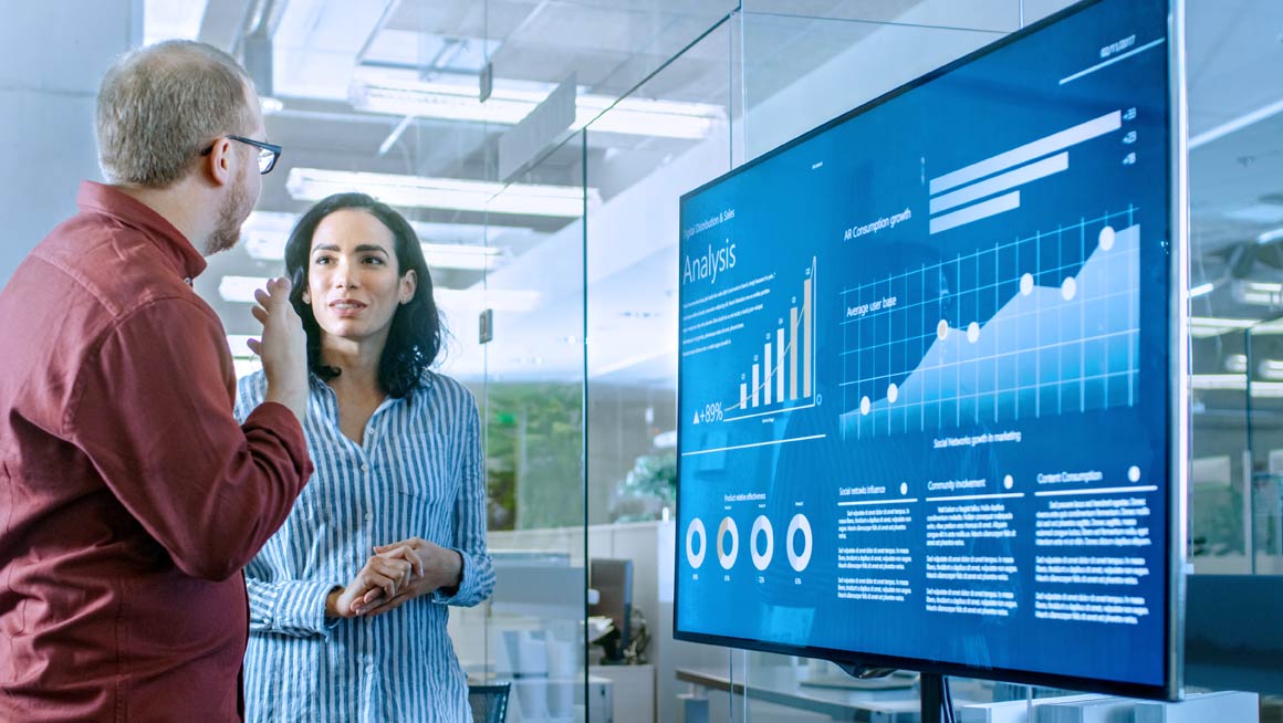 A man and a woman have a discussion in front of a monitor holding data.
