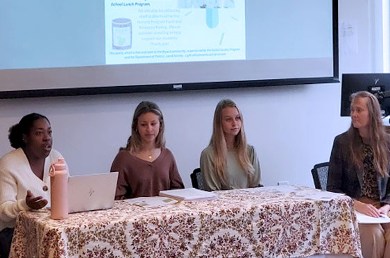 Bryant University Professor Nicole Frenier sits a table with three students giving a presentation.
