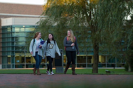 A group of three female students walk along a brick pathway in front of the George E. Bello Center for Information and Technology at Bryant University.