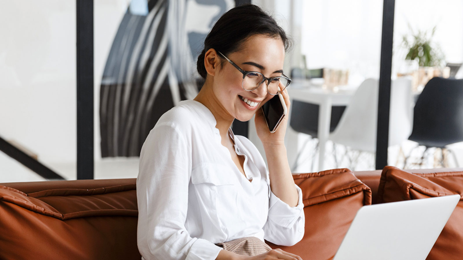 A woman with glasses sitting on the couch on the phone with her laptop.