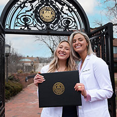 Two Bryant physician assistant graduates stand with a diploma in front of the archway.